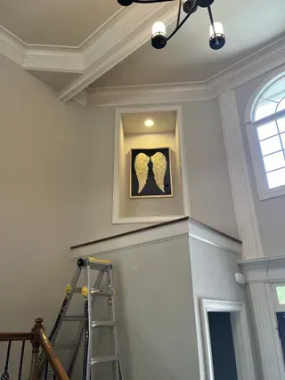 A framed print of golden angel wings is centered in an illuminated wall niche above a stairwell with a metal ladder nearby.