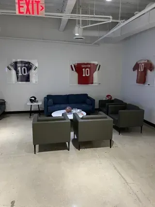 A lounge area with three framed jerseys, two chairs, a blue couch, and a small table on a light-colored concrete floor.