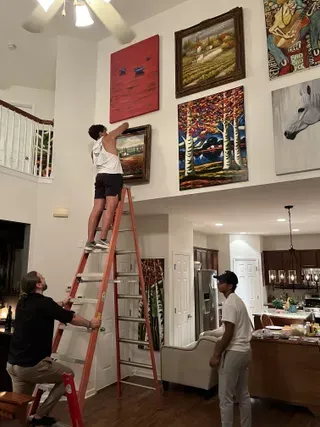 Three people work to hang several large paintings on a high wall in a spacious home with an orange ladder.