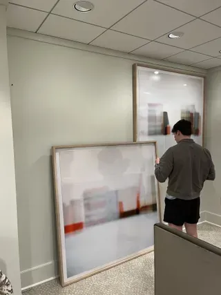A person stands in a room, holding a large, framed abstract painting while another identical painting hangs on the wall.