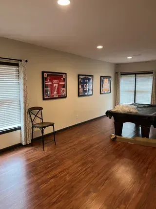 A room with light walls, hardwood floors, a single chair, three framed sports jerseys on the wall, and a pool table.