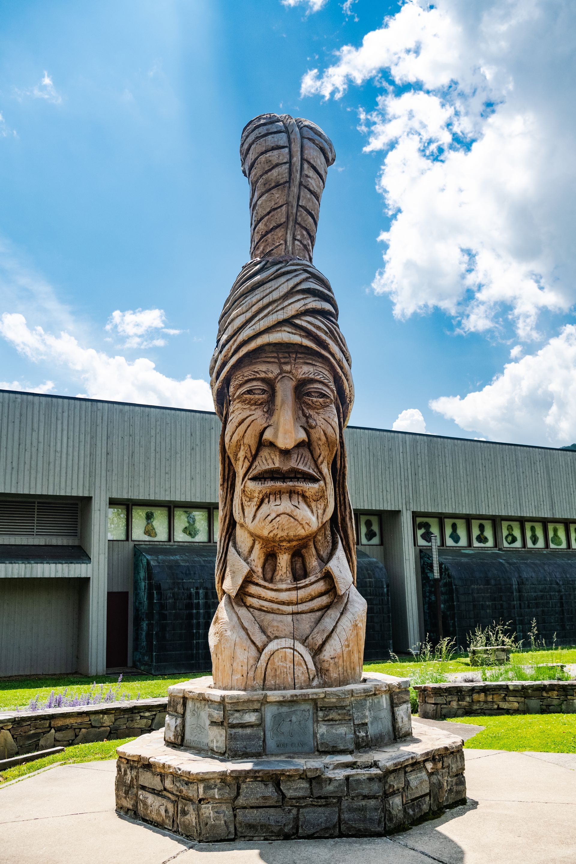 Carved wooden Native American statue on a stone base, outdoors, blue sky.