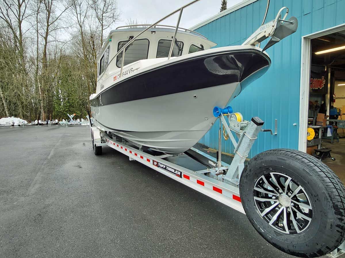 Boat on a trailer in front of a blue building, gray boat with black trim, spare tire mounted on the trailer.