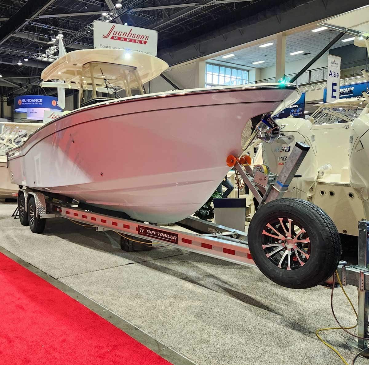 White boat on a trailer, displayed at a boat show with red carpet, neutral background.