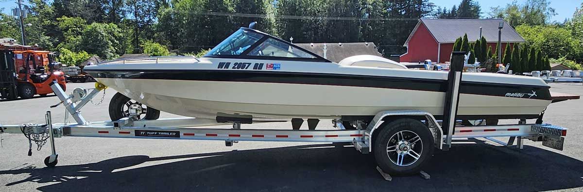 A white speedboat on a trailer in a paved lot, with trees and a building in the background.