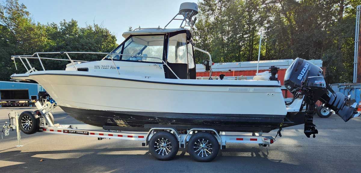 White boat on a trailer, with a blue motor, parked on asphalt with trees in the background.