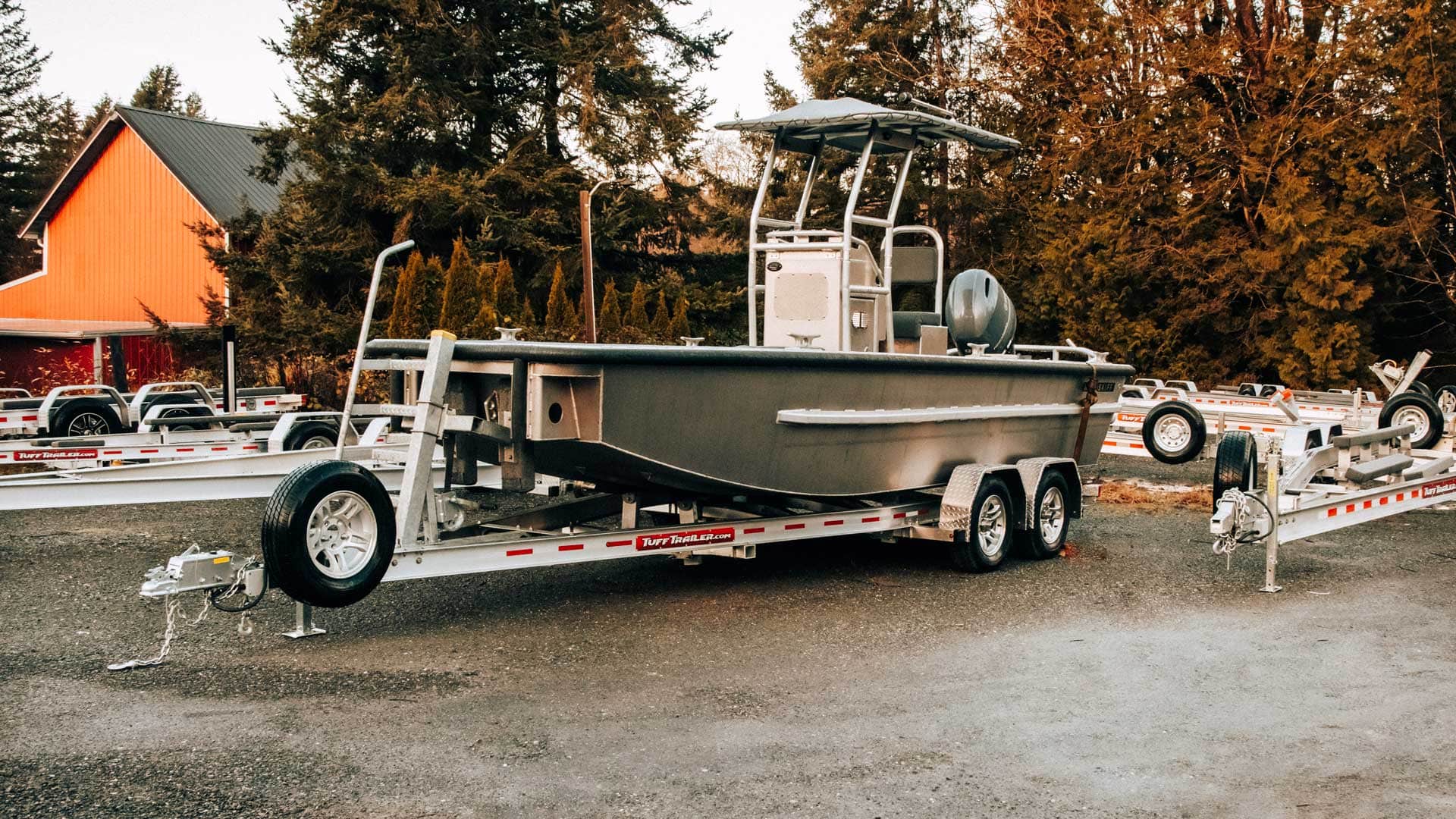 Gray aluminum boat on a trailer, with a barn and trees in the background.