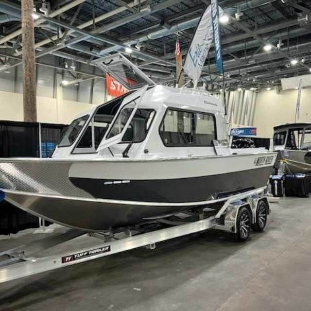 Gray and white aluminum boat on a trailer, in a convention hall, with a raised roof and windows.