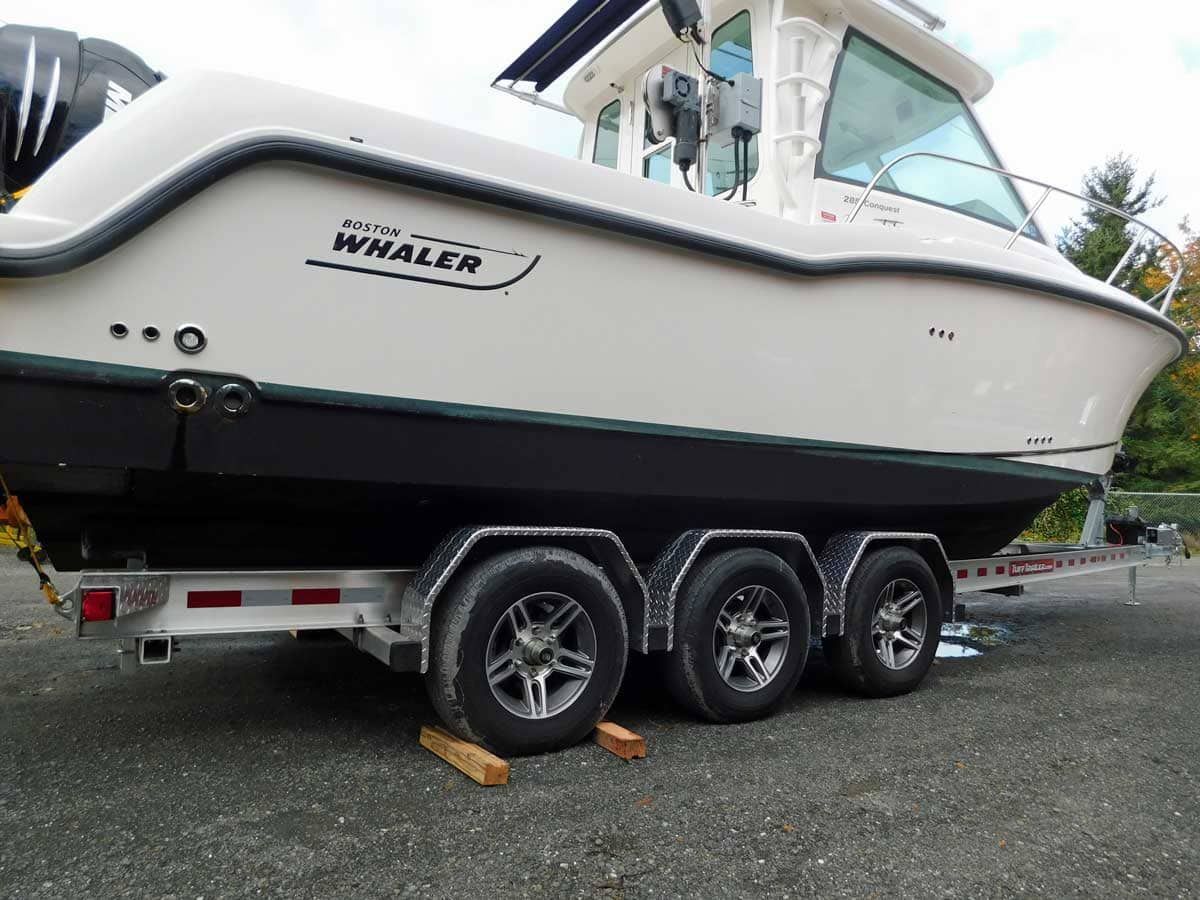 White Boston Whaler boat on a triple-axle trailer, sitting on wooden blocks, in a gravel lot.