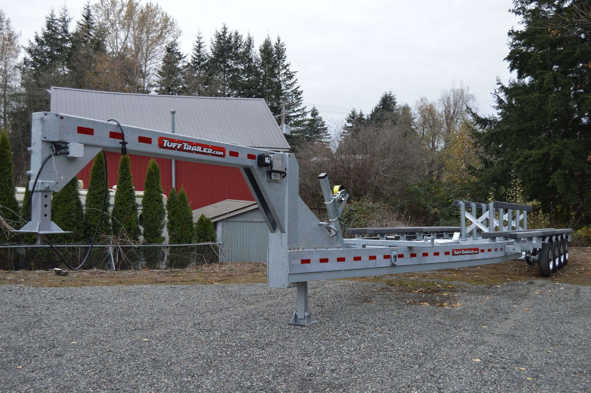 A long, gray heavy-duty trailer parked on gravel in front of a red barn and trees.
