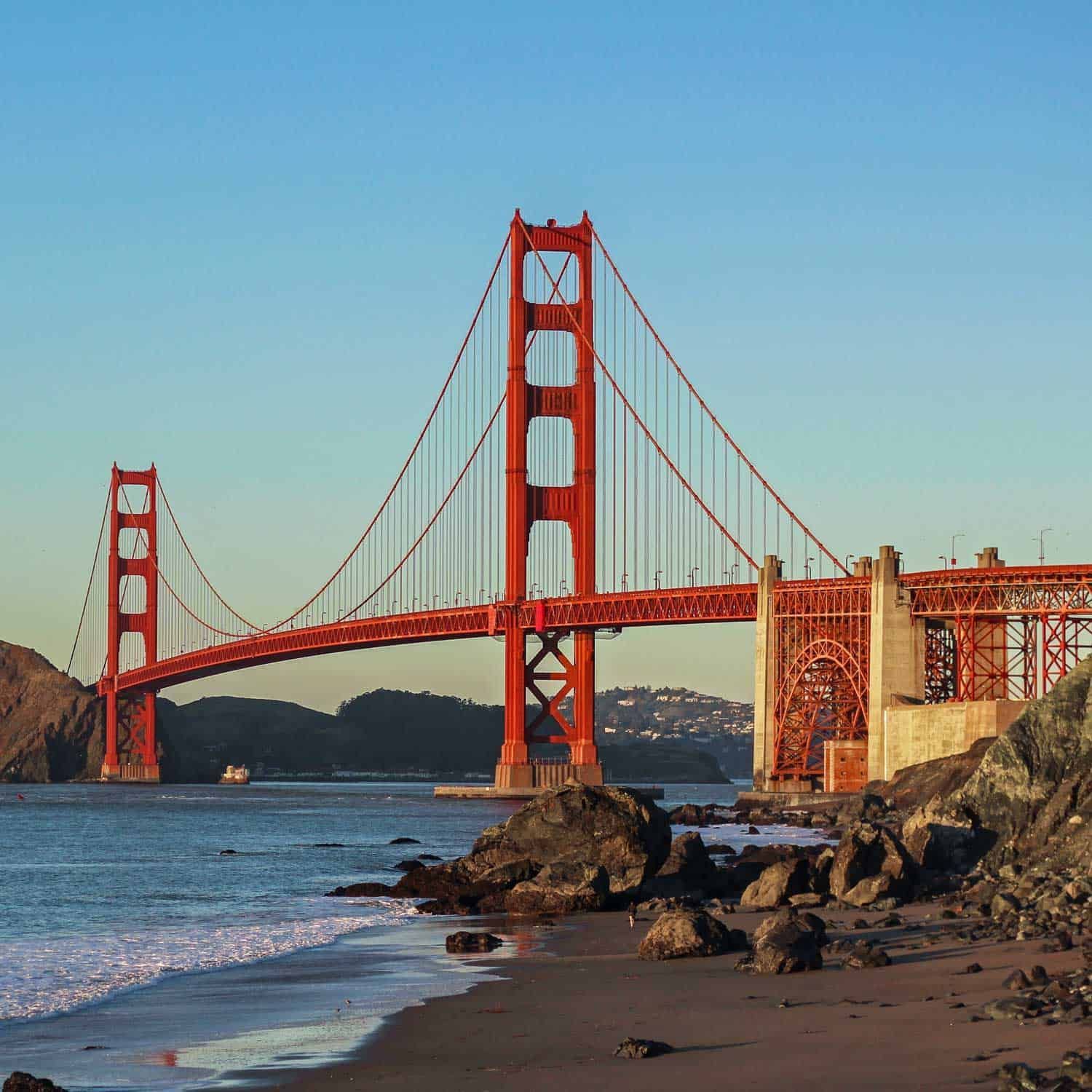 Golden Gate Bridge spans the water, towers and deck in reddish-orange; ocean in foreground under blue sky.
