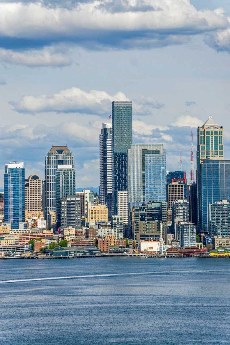 Seattle skyline on a sunny day, with a body of water in the foreground and a cloudy sky.