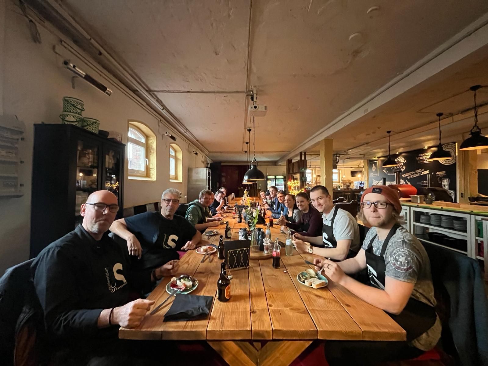In einem gemütlichen Restaurant sitzen Menschen an einem langen Holztisch, unterhalten sich und essen gemeinsam.