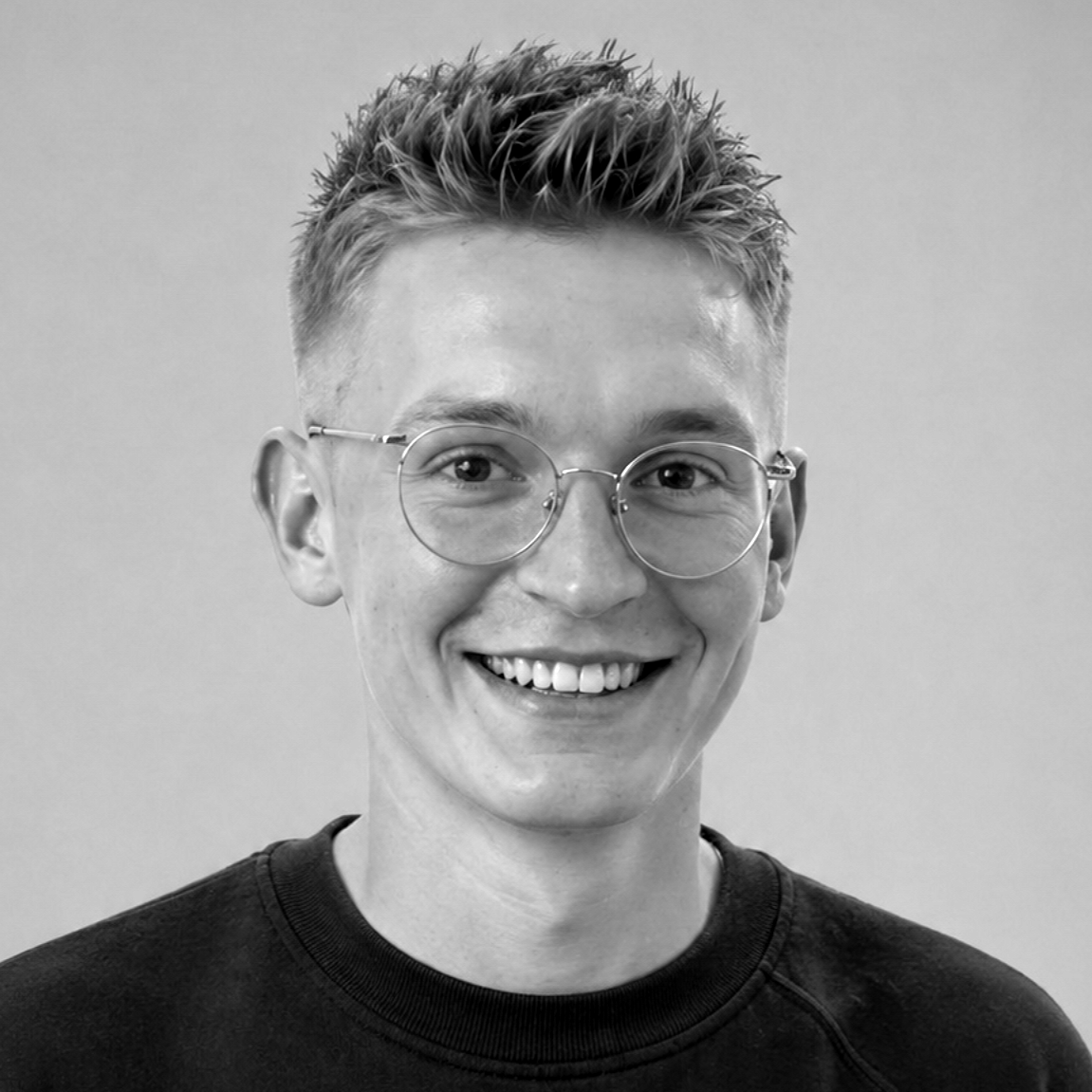 Lächelnde Person mit Brille und schwarzem Sweatshirt vor grauem Hintergrund