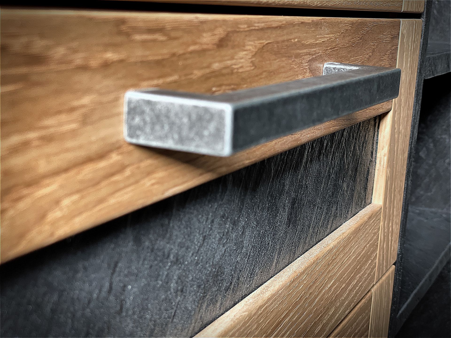 Nahaufnahme eines metallenen Schubladengriffs an einem Holzschrank; die Schublade ist mit dunklem Stoff ausgekleidet.