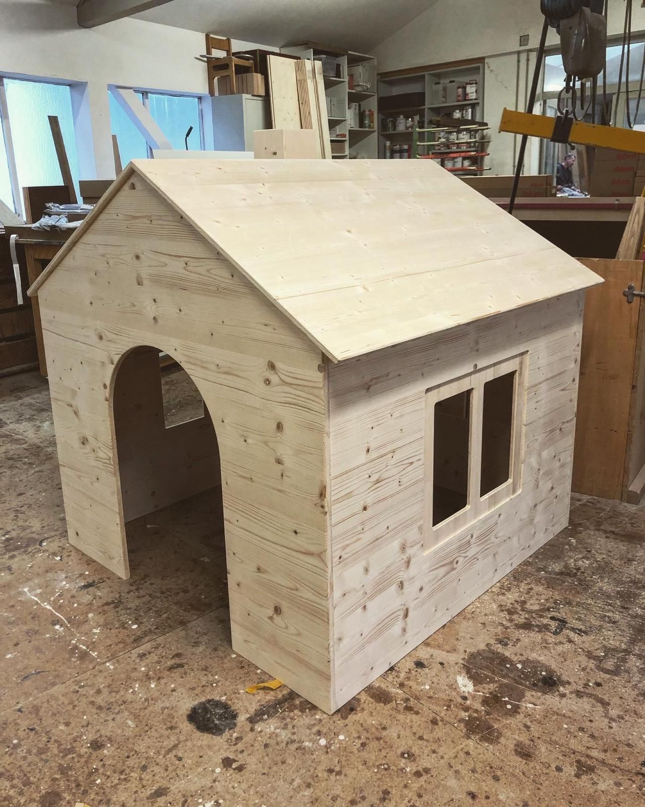 Kleines, unfertiges Holzspielhaus mit Satteldach und Fenster in einer Werkstatt.