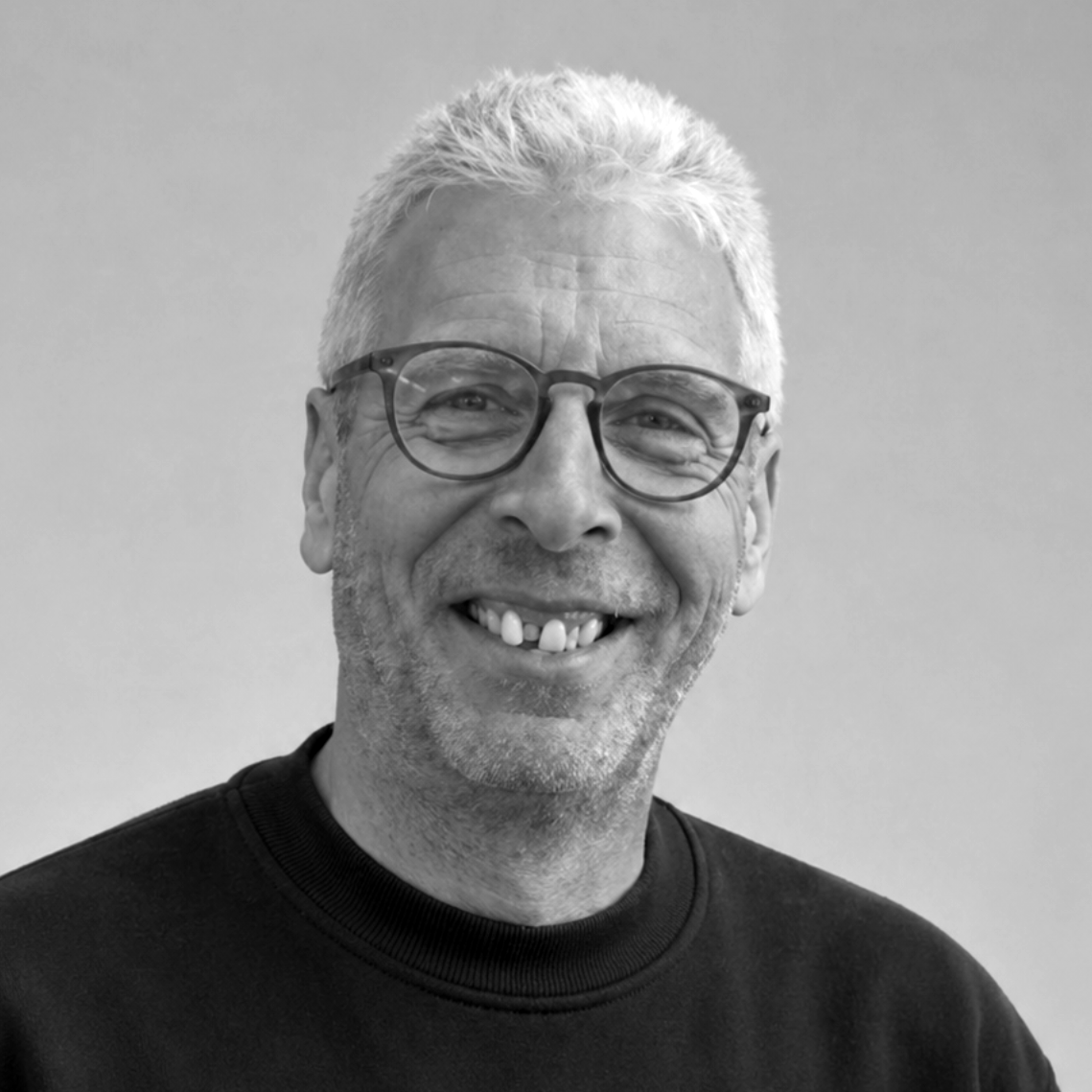 Lächelnder Mann mit Brille und grauem Haar, der einen schwarzen Pullover trägt, vor einem schlichten grauen Hintergrund