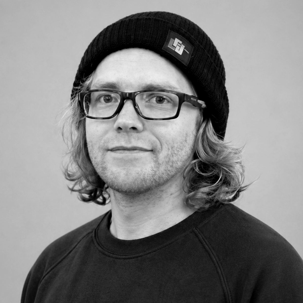 Person mit schwarzer Mütze und Brille vor grauem Hintergrund, die in die Kamera schaut.