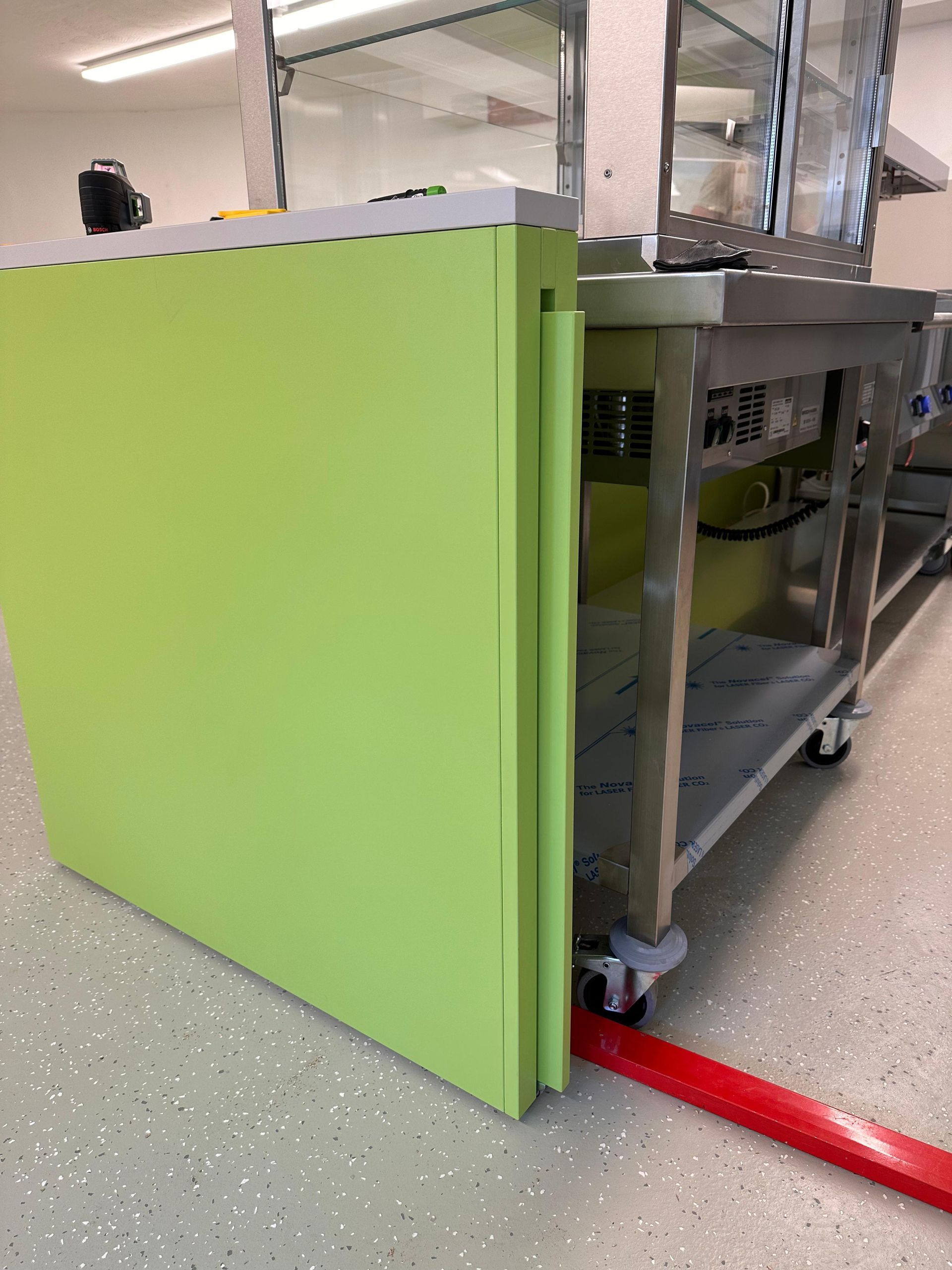 Ein leuchtend grüner Wagen oder Schrank neben einem Edelstahltisch in einer Werkstatt, mit einem roten Gurt auf dem Boden.