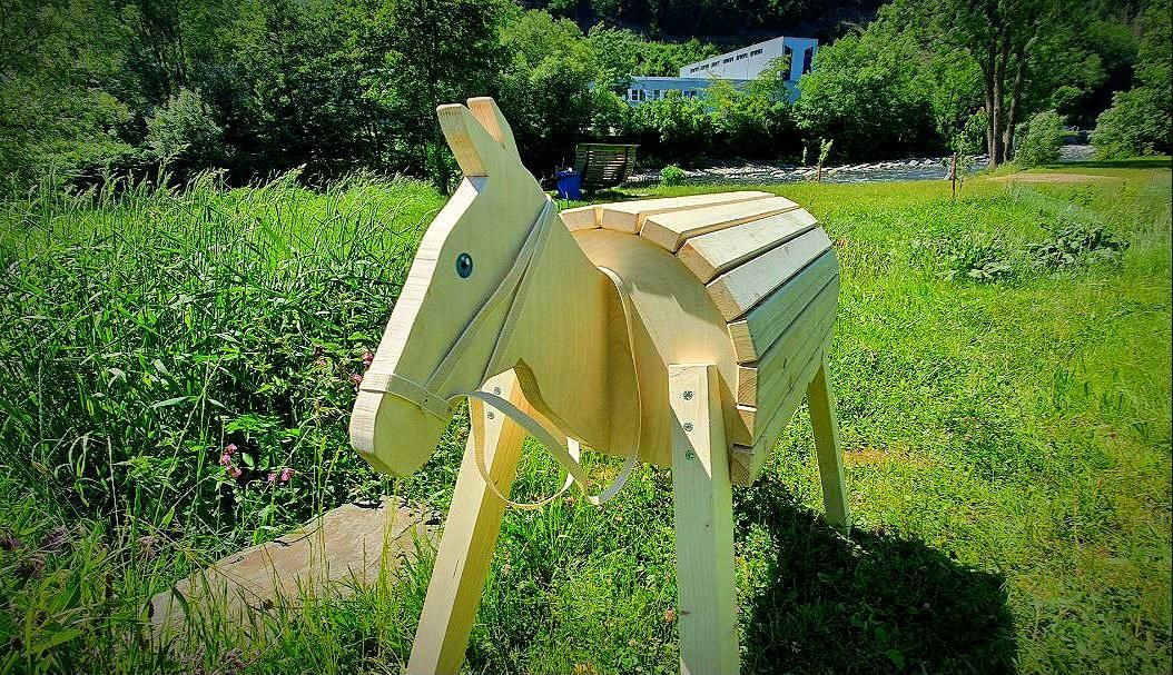 Holzpferdeskulptur auf einer Wiese mit Bäumen und Blumen im Hintergrund
