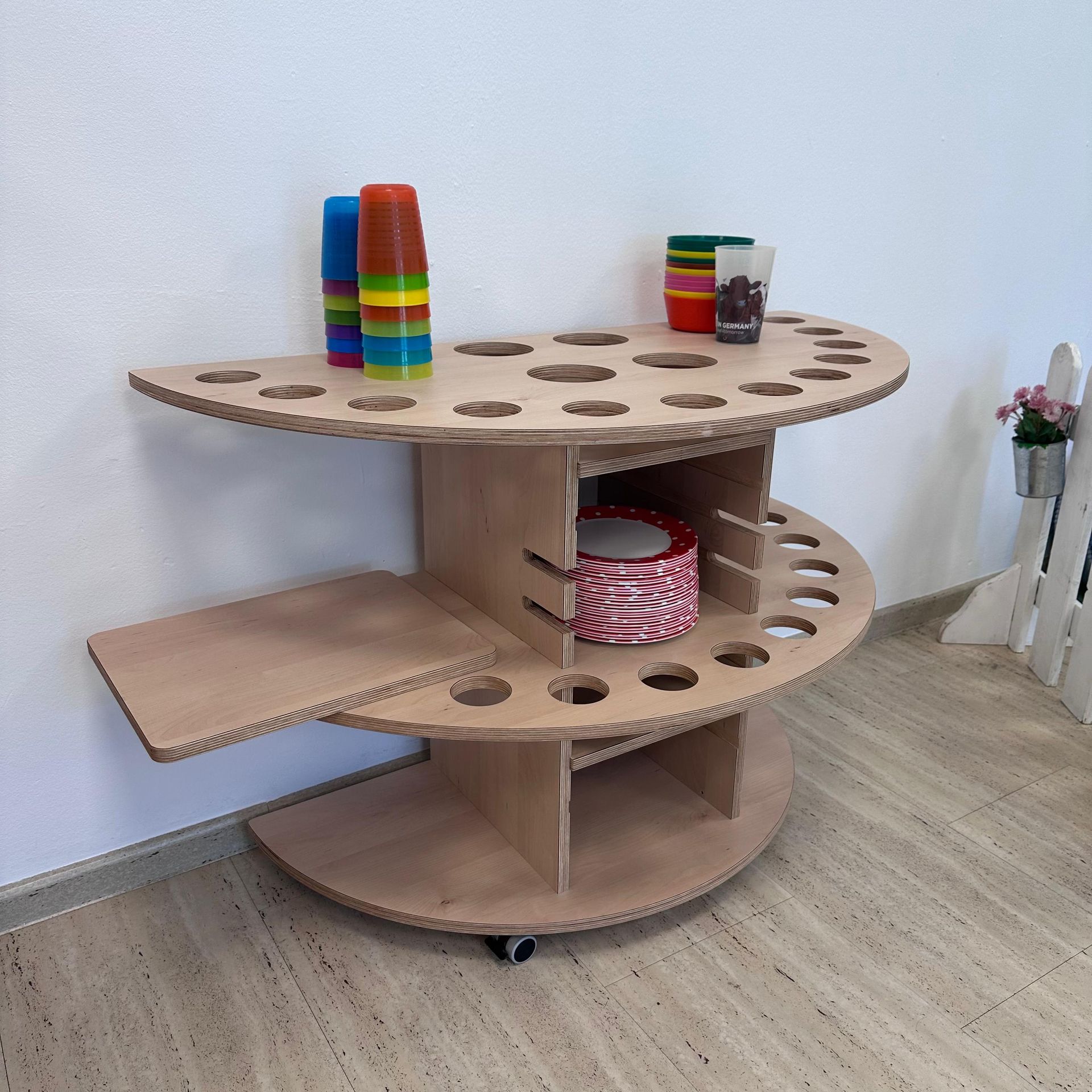 Sensorische Spielstation aus Holz mit bunten Stapelbechern, einem Seitenregal und Körben auf Rollen vor einer weißen Wand