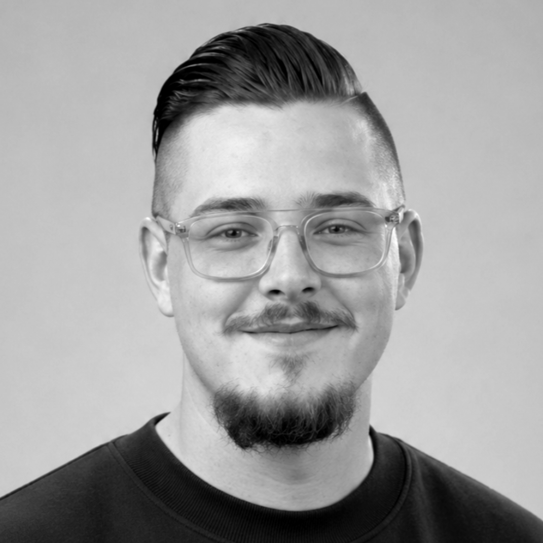 Person mit Brille und Schnurrbart, die ein schwarzes Sweatshirt trägt, vor grauem Hintergrund.