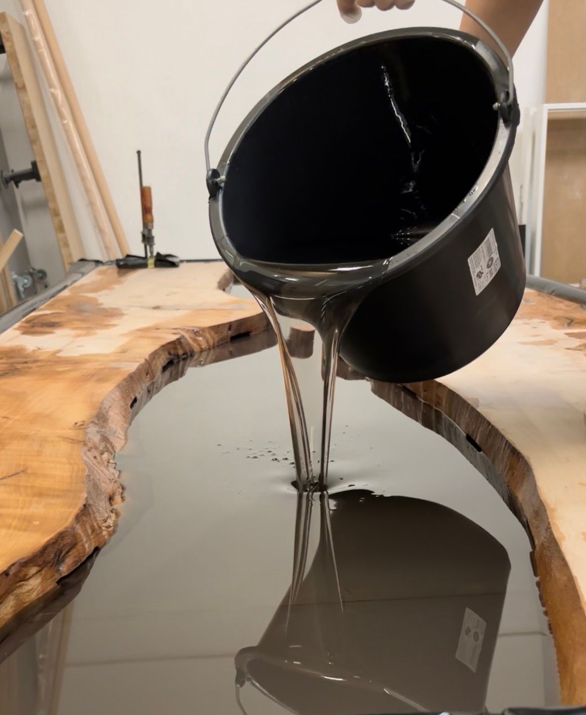 Ein schwarzer Eimer gießt glänzende Flüssigkeit in eine holzförmige Form auf einem Werkstatttisch.