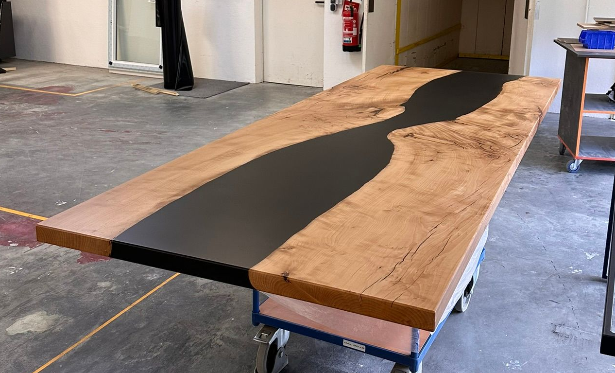 Lange Holzplatte auf blauen Sägeböcken in einer Werkstatt, mit einer dunklen, mit Epoxidharz gefüllten Mittelrille.