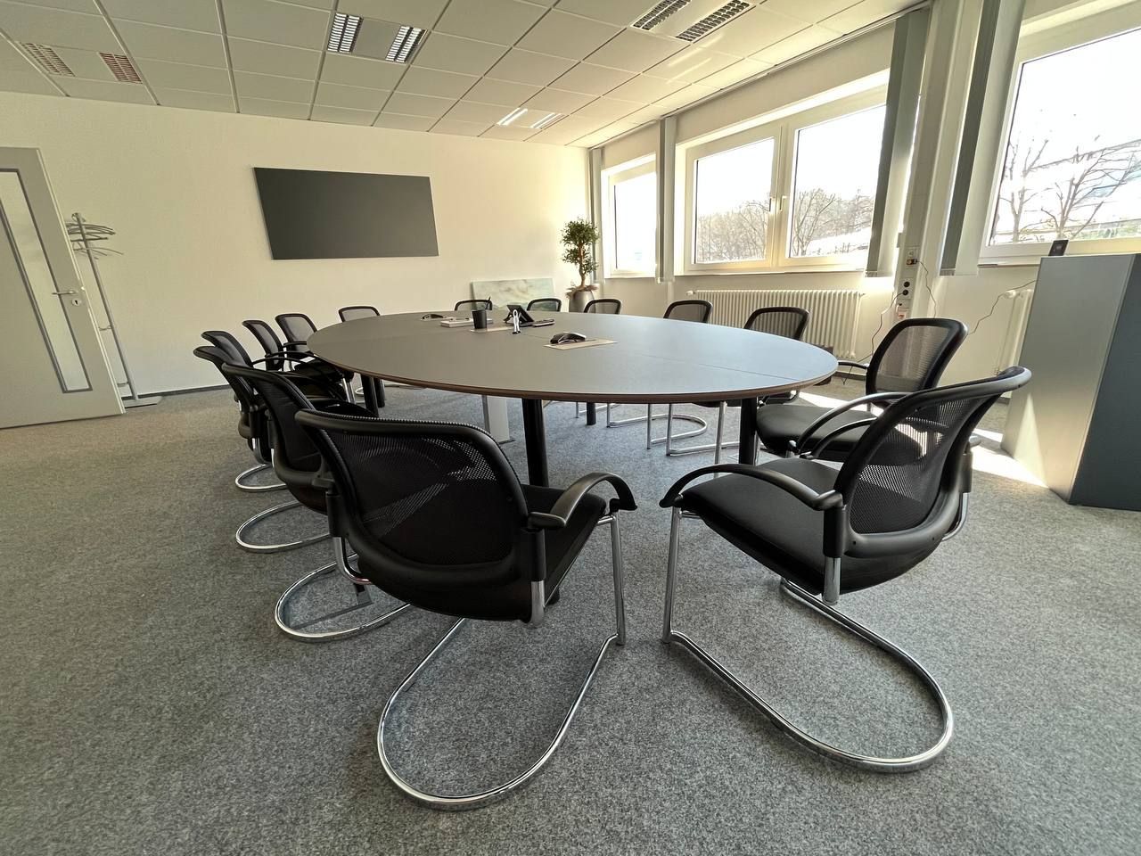 Konferenzraum mit rundem Tisch, schwarzen Stühlen, an der Wand montiertem Fernseher und Fenstern, die Tageslicht hereinlassen