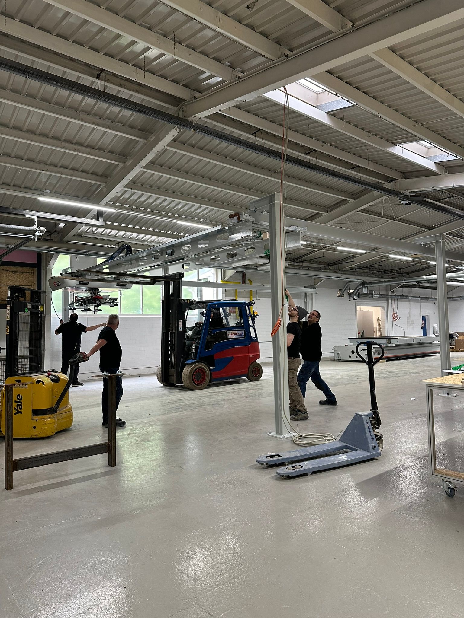 Arbeiter in einer Fabrikhalle mit einem gelben Gabelstapler, einer roten Maschine und Oberlichtern.