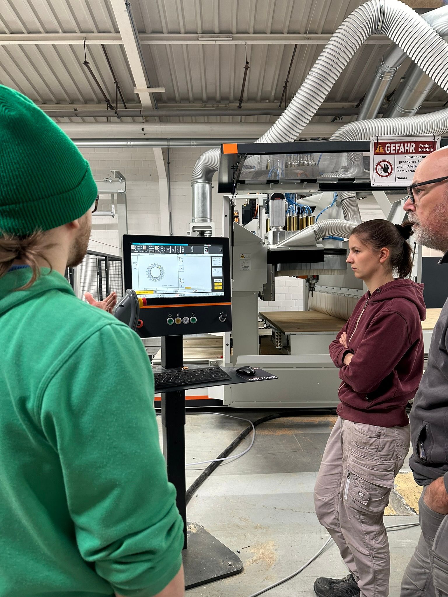 Zwei Personen in einer Fabrik schauen auf einen Maschinensteuerungsbildschirm; über ihnen befinden sich Lüftungsschlitze und Industrieanlagen.