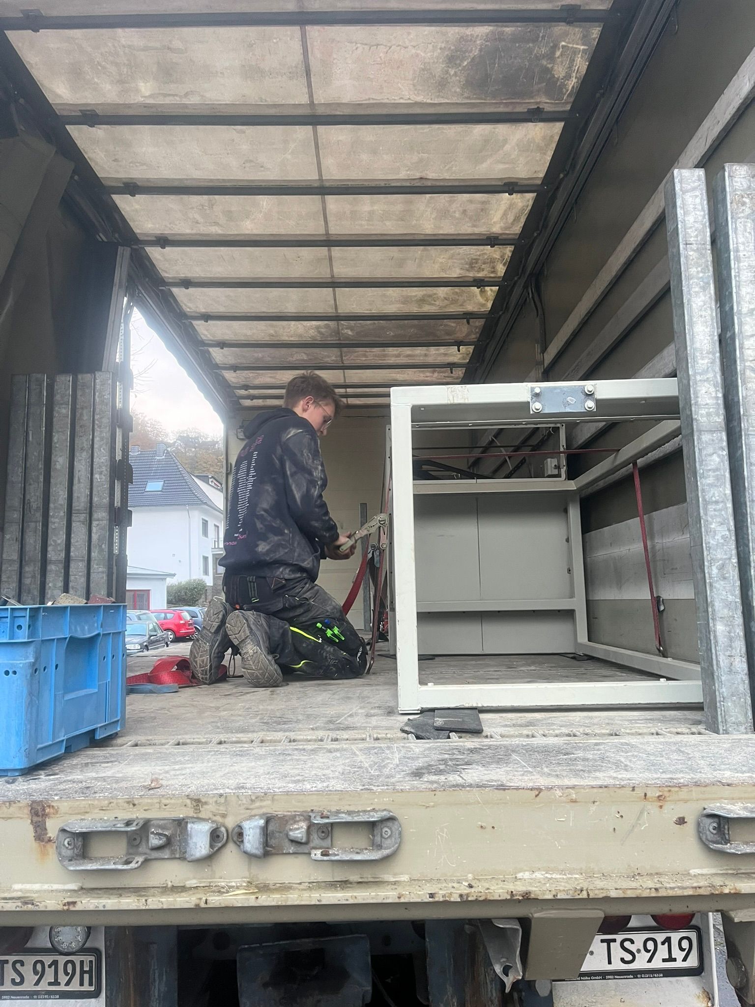 Eine Person kniet auf der Ladefläche eines Lastwagens neben einem weißen Schrank und verlädt oder sichert Ausrüstung unter einem Vordach.