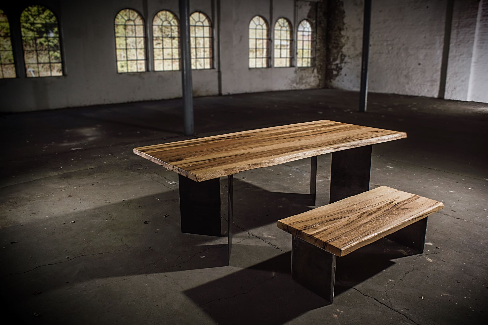 Rustikaler Holztisch und Bank in einem dunklen, leeren Industrieraum mit Bogenfenstern.
