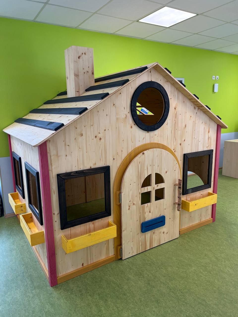 Spielhaus aus Holz mit Spitzdach, Fenstern und Bogentür in einem hellgrünen Innenraum Kindergarten

