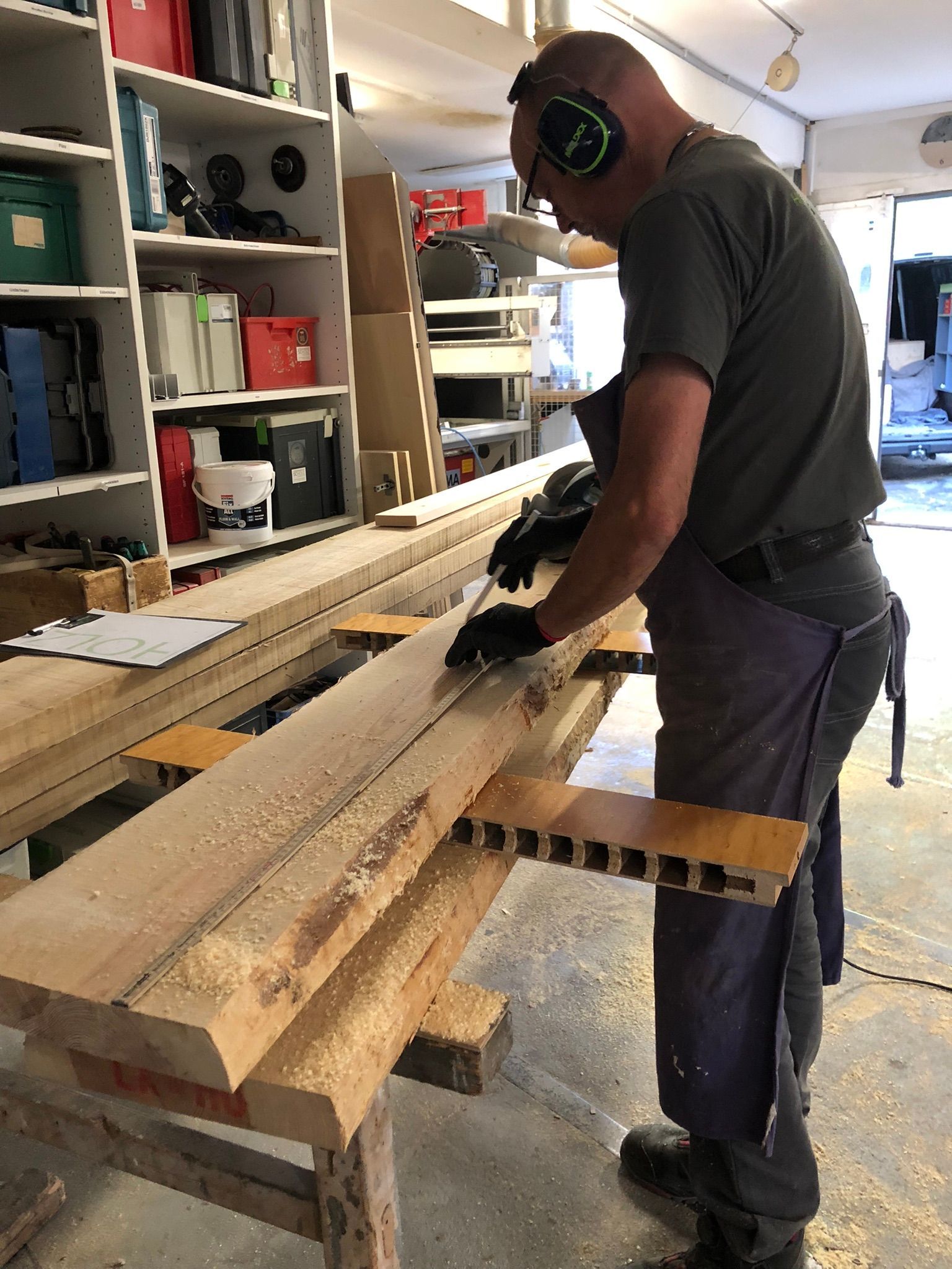 Eine Person schleift an einem Werkstatttisch ein Holzbrett; sie trägt Gehörschutz und Handschuhe.