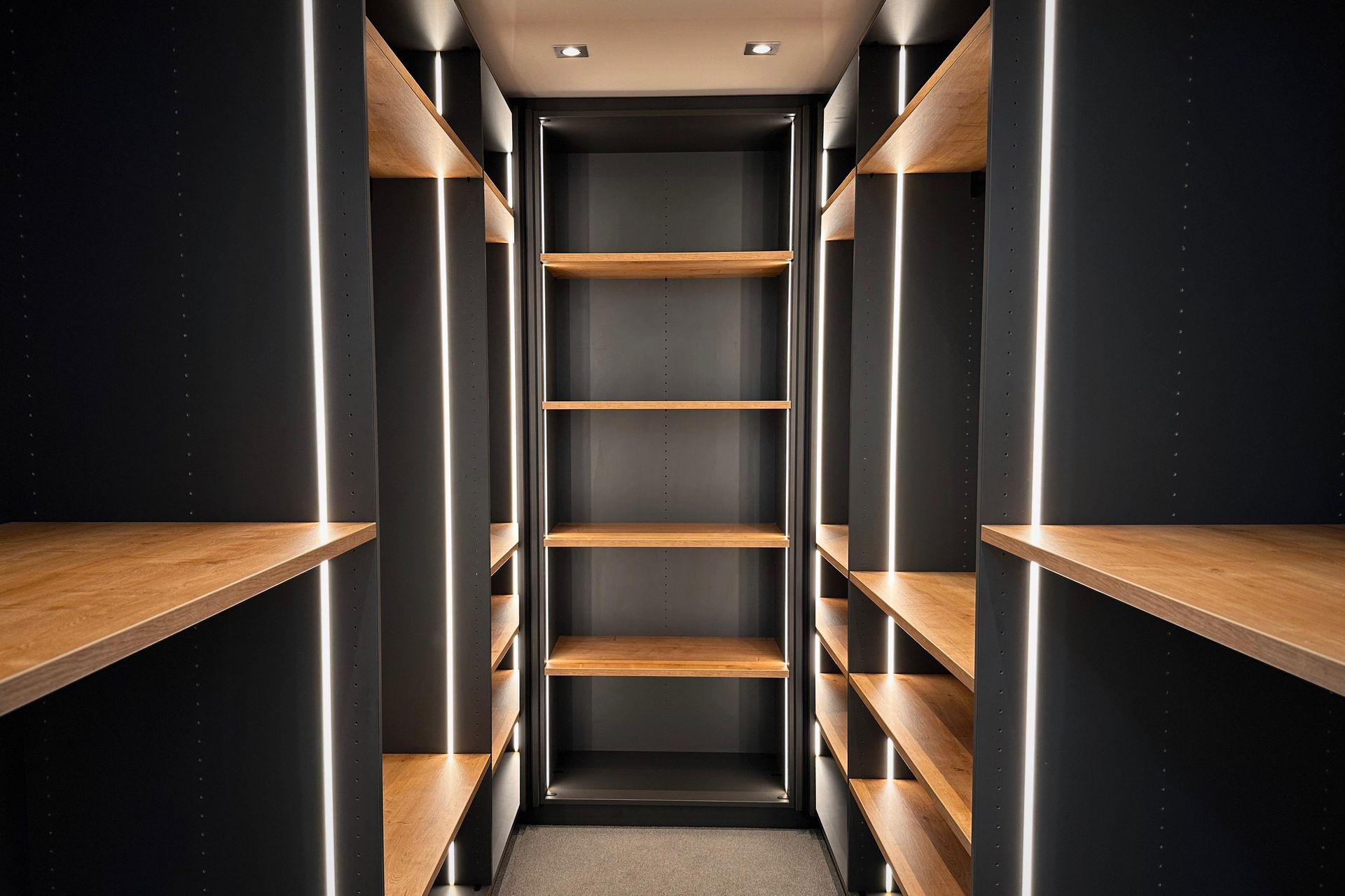 Schmaler begehbarer Kleiderschrank mit dunklen Regalen, Holzelementen und warmem Einbaulicht