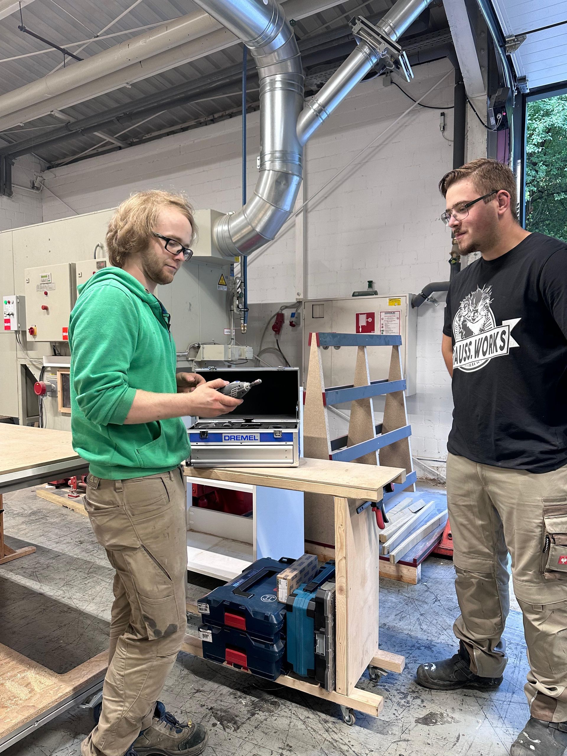 Zwei Personen stehen an einer Werkbank in einer Werkstatt, eine hält ein Werkzeug in der Nähe eines 3D-Druckers und eines Kartons.