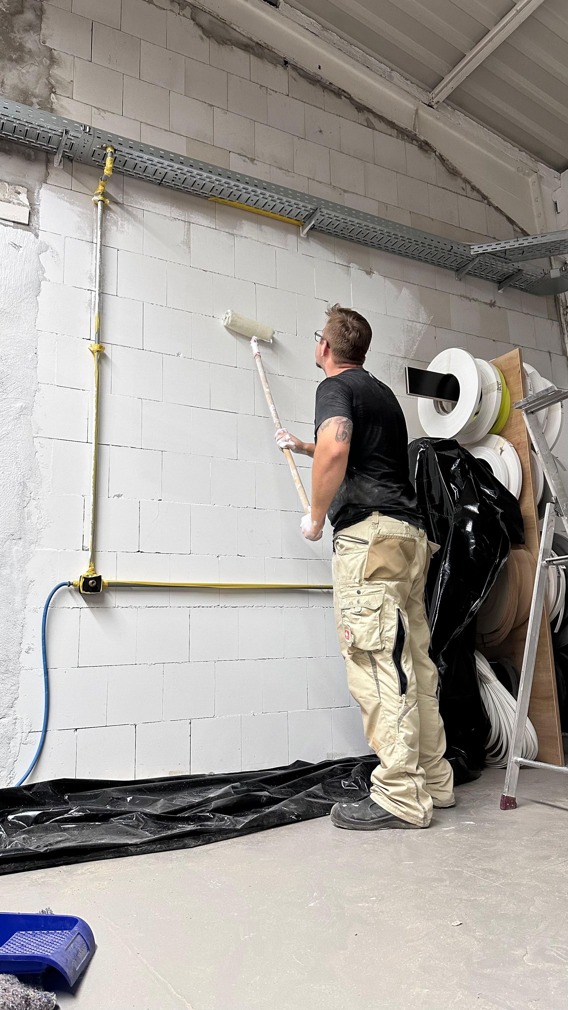 Ein Arbeiter installiert in einem unfertigen Raum Kabel an einer Betonsteinwand, mit Kabelrollen und Leitungsrohren.