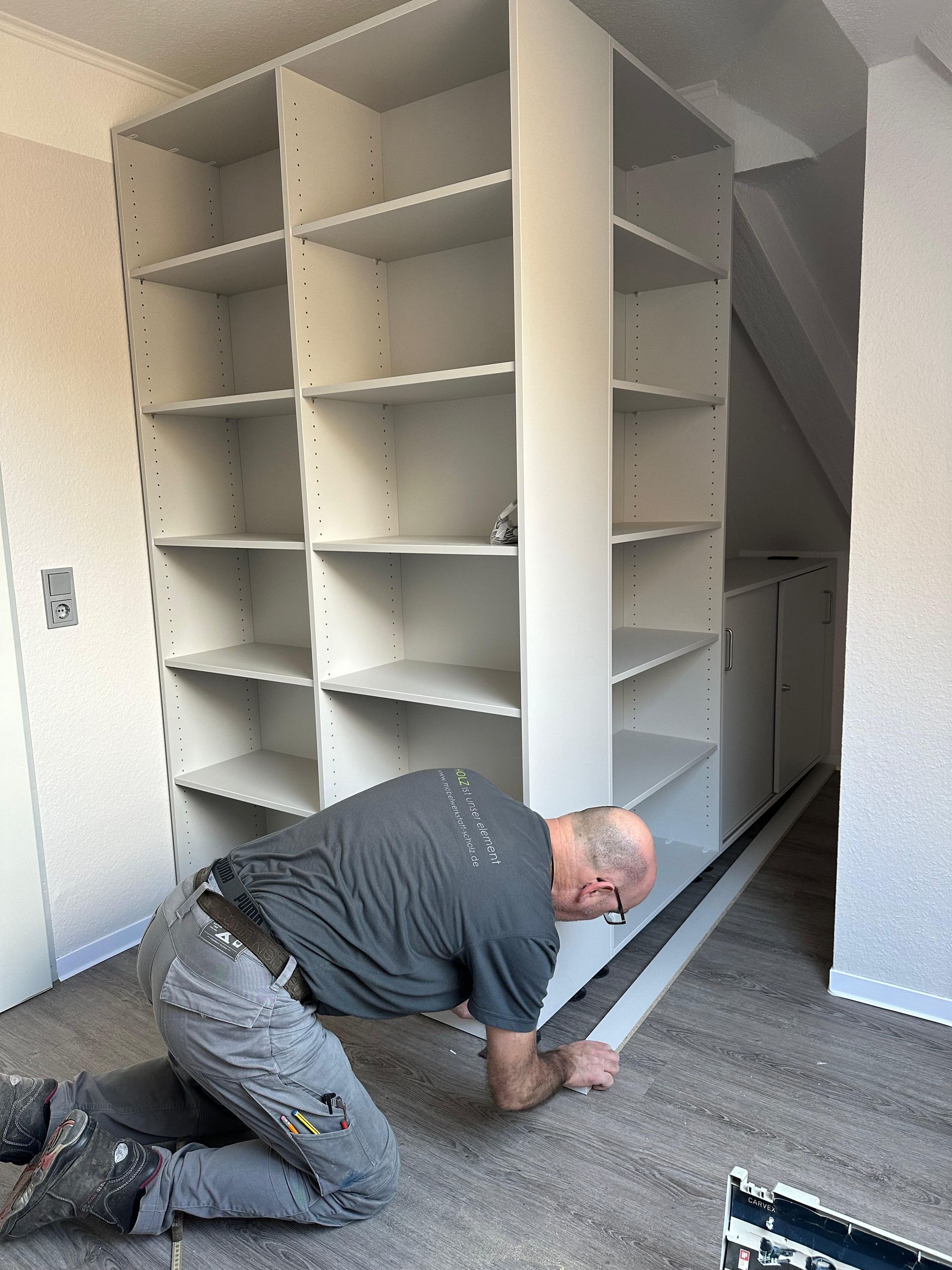 Eine Person kniet neben neuen weißen Regalen in einem Zimmer und misst den Boden mit einem Maßband.