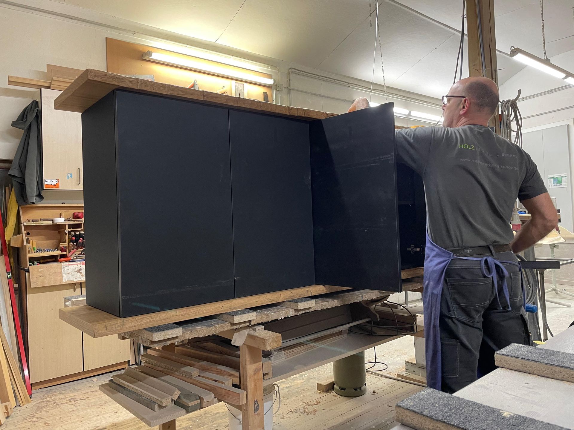 Ein Mann schleift in einer Werkstatt einen großen schwarzen Schrank ab; um ihn herum liegen Holzbretter und Werkzeuge.