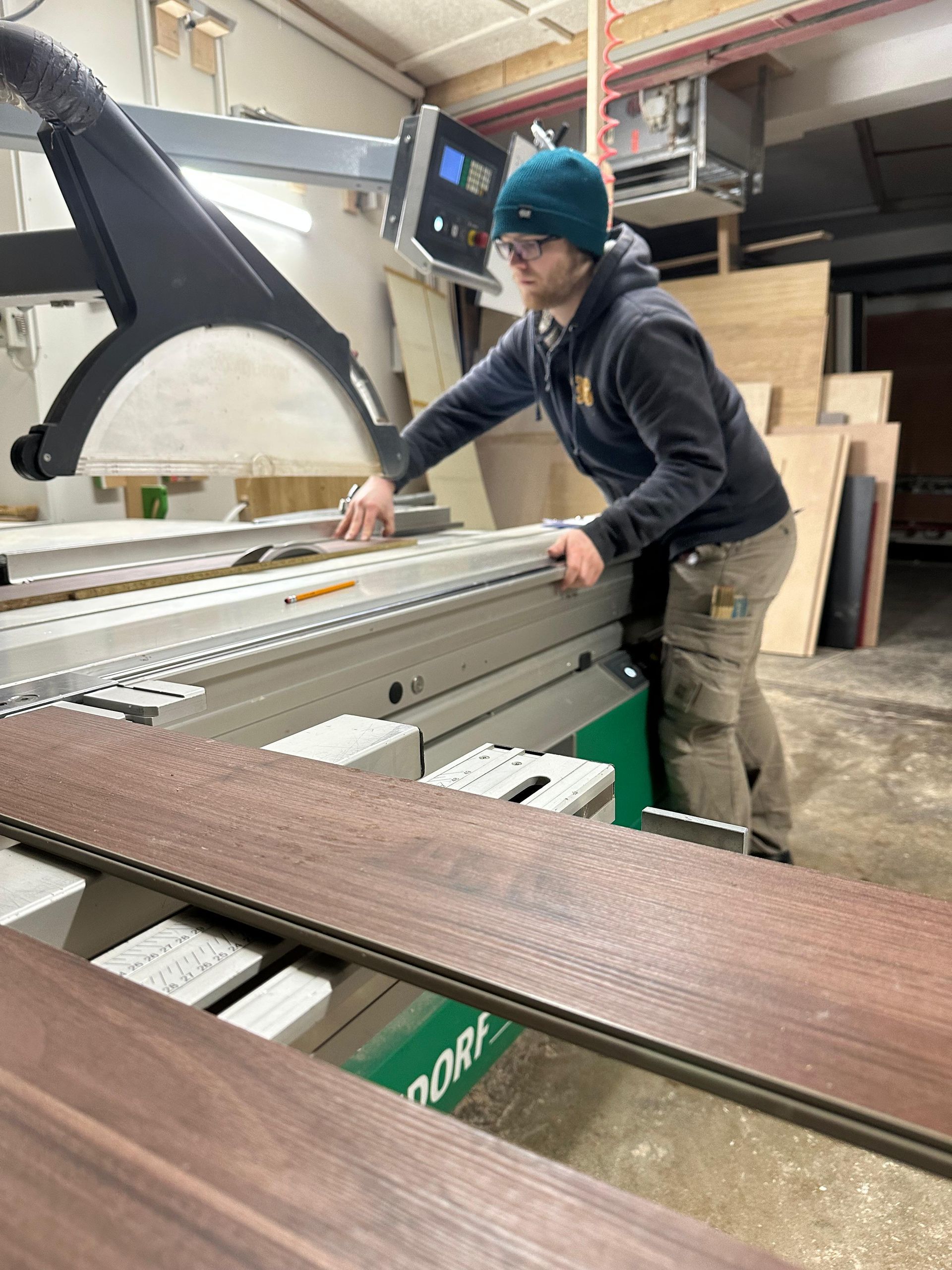 Ein Arbeiter bedient in einer Werkstatt eine große Holzbearbeitungsmaschine; auf dem Tisch liegen Bretter.