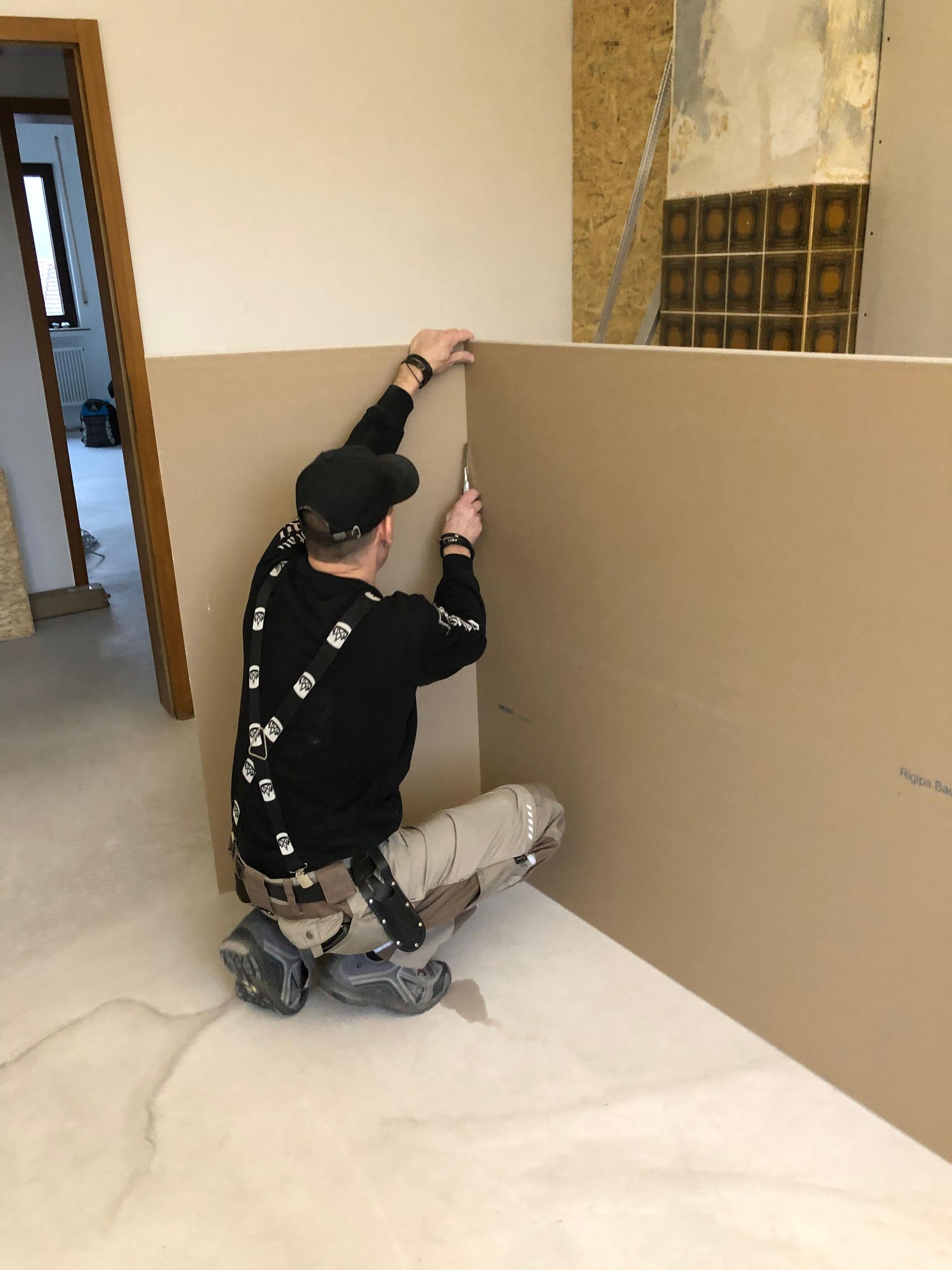 Ein Arbeiter kniet, um eine beige Wand in einem Raum zu streichen, der gerade renoviert wird.
