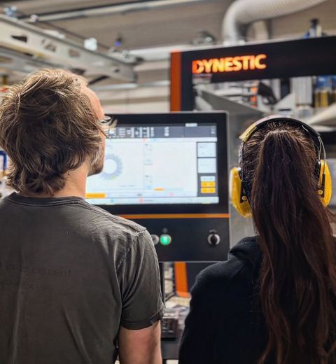 Zwei Arbeiter mit Gehörschutz überwachen einen Maschinenkontrollbildschirm in einer Fabrik.