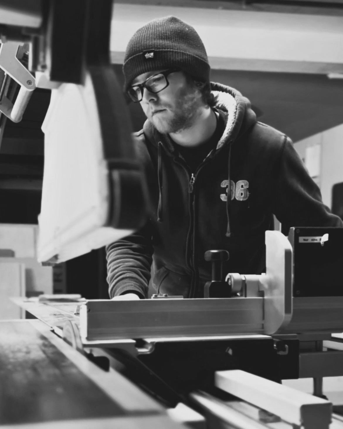Eine Person mit Mütze und Brille arbeitet an einer Holzbearbeitungsmaschine in einer Werkstatt.