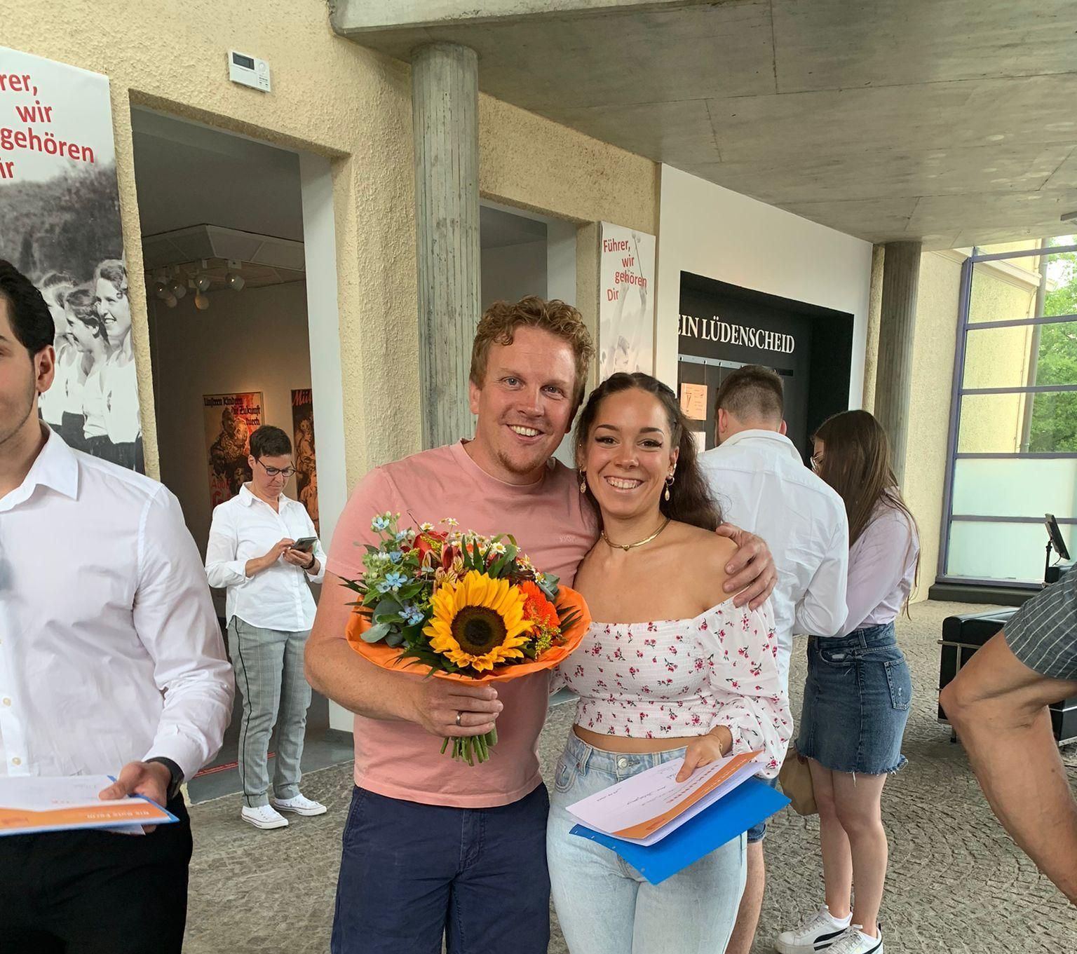 Zwei lächelnde Personen halten Blumen und Urkunden bei einer Abschlussfeier in den Händen.