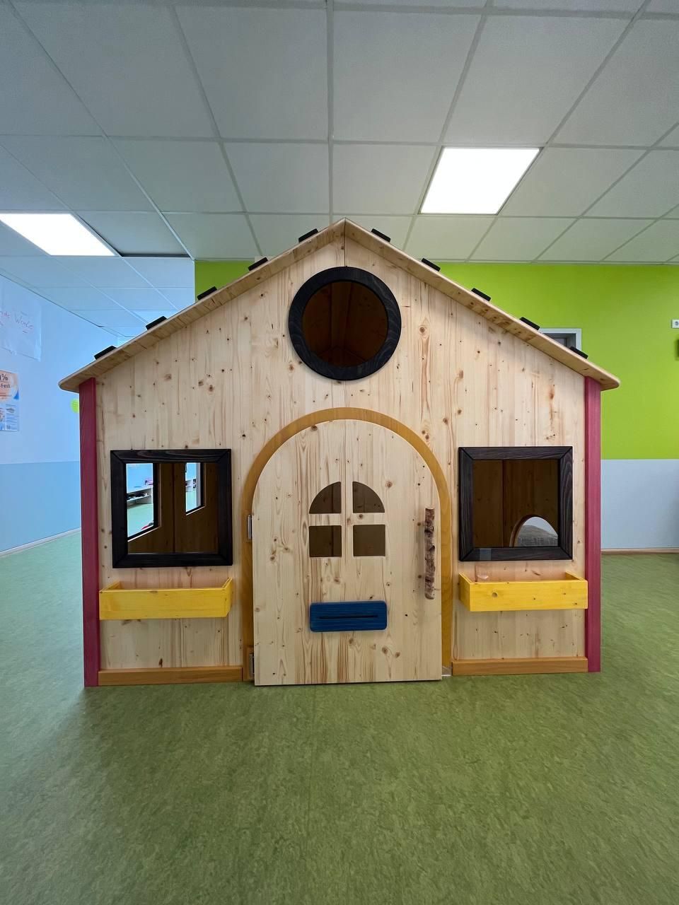 Kleines Holzspielhaus mit Fenstern und blauer Tür in einem farbenfrohen Indoor-Spielzimmer