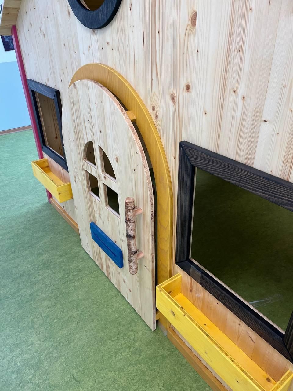 Kleine hölzerne Spielhausmauer mit gewölbter weißer Tür, schwarz gerahmten Fenstern und gelber Umrandung auf grünem Rasen