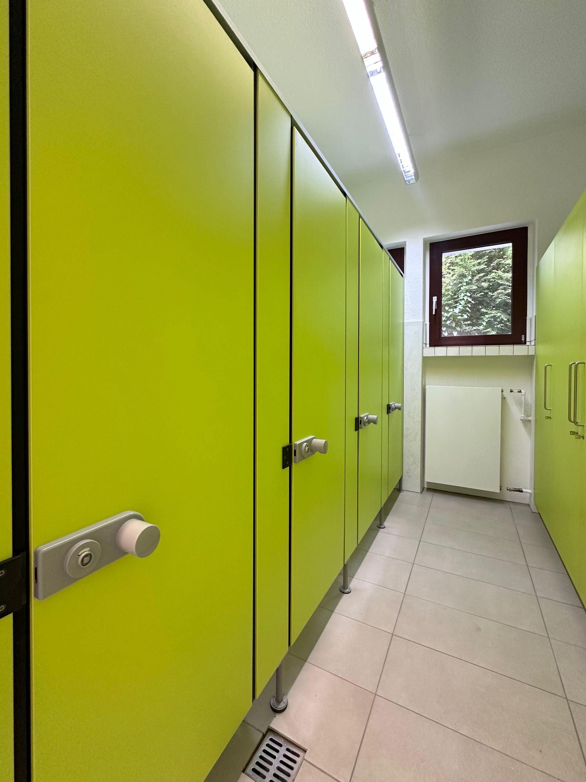 Hellgrüne Toilettenkabinen in einem schmalen, gefliesten Flur mit Neonbeleuchtung und einem Fenster am Ende.