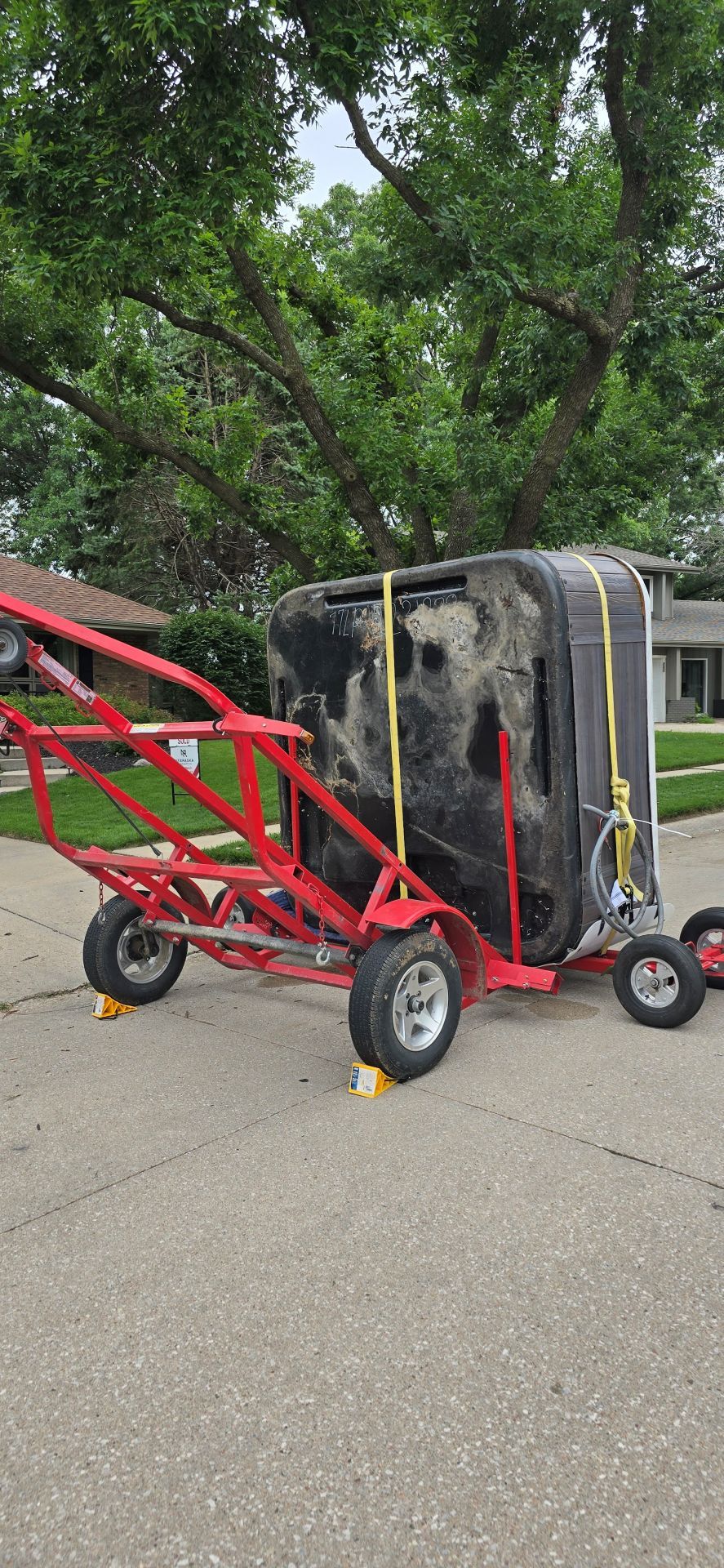 Red utility cart with large wheels and a black crate, outdoors on a paved lot with trees behind.