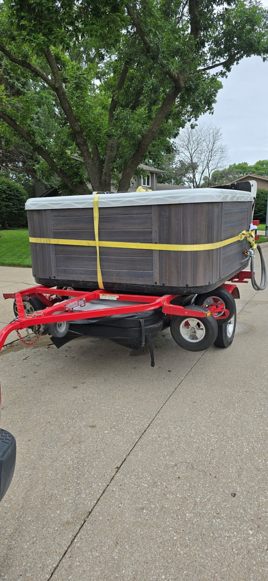 Red trailer carrying a large gray wrapped load secured with yellow straps on a street under trees